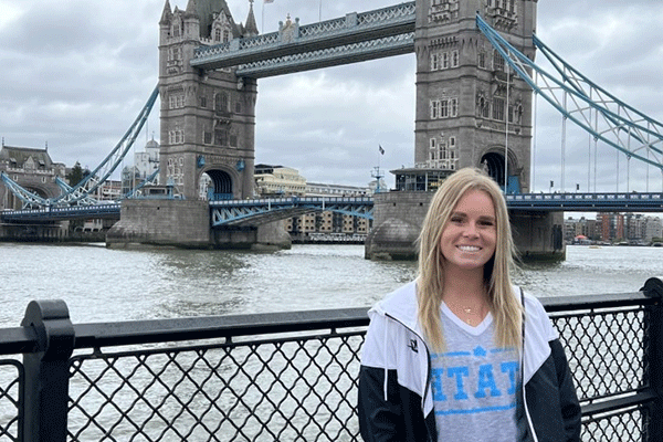 A female student with blonde hair poses in front of the London Bridge, a bridge with two stone towers. Water is behind her. She wears a grey-and-blue STATE T-shirt and a black-and-white jacket.