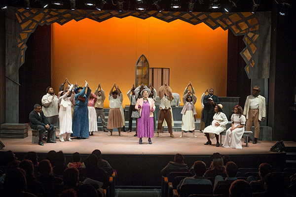 A theater performance on a stage. The cast includes 20+ Black performers wearing a variety of early 20th-century clothing. The stage is decorated with a dark orange stained-glass window in the back, a wardrobe changing divider, benches, and other orange stained-glass hanging from the ceiling. Several actors are visible with their arms raised in prayer. An audience sits in seats in the theater. 